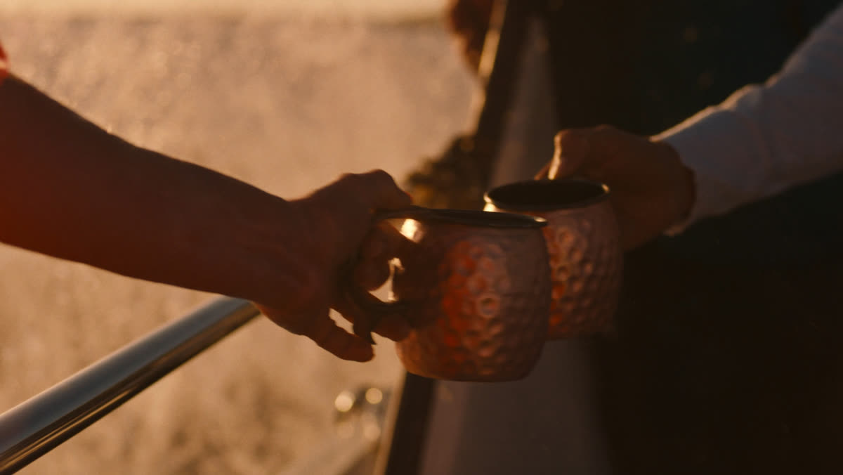 Two hands clinking copper mule mugs over the golden wake of a boat at sunset