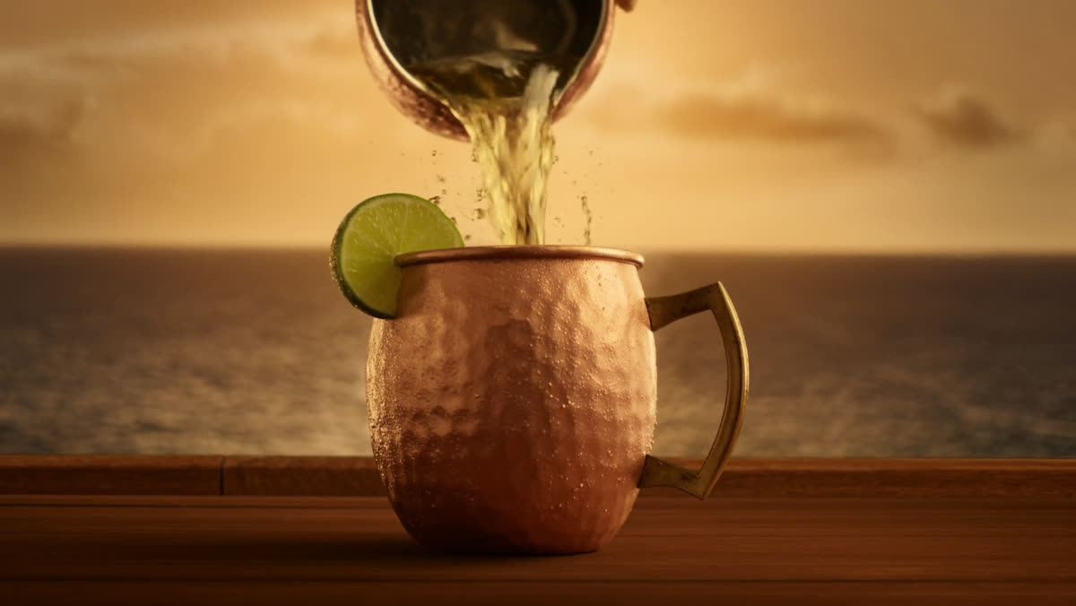 Ginger beer pouring into a frosted copper mule mug with a lime wheel, ocean sunset behind